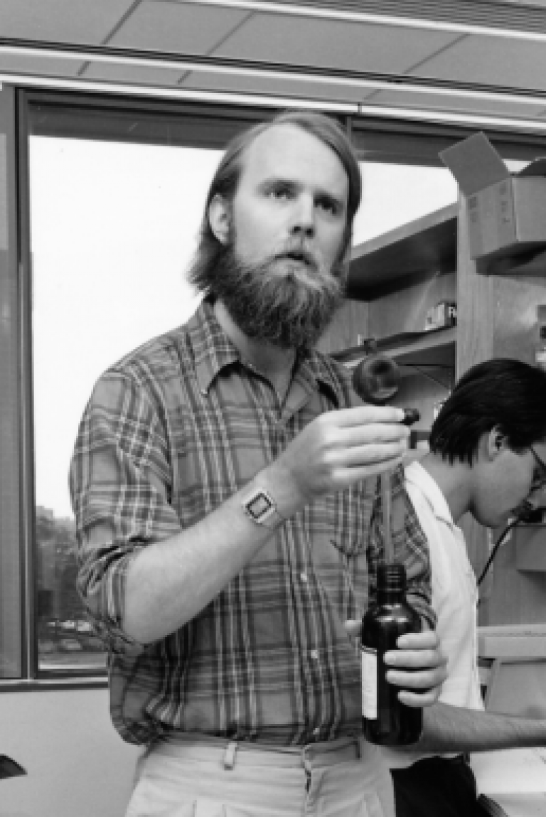 A man in a lab talks while holding a bottle of reagent.